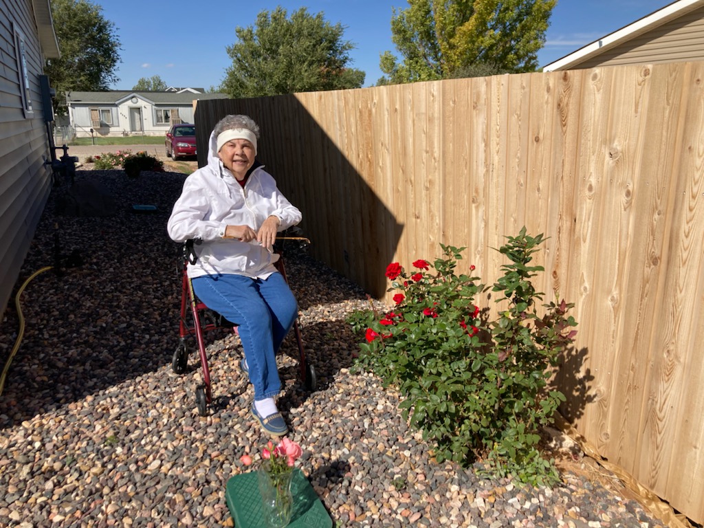 An elderly woman poses in a garden with bloomed flowers.