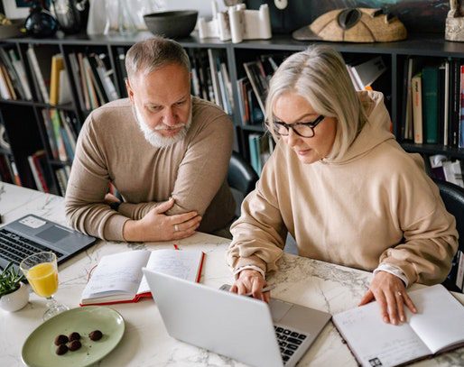 A busy elderly man and woman working on their laptops on a table with bookshelves in the background.
