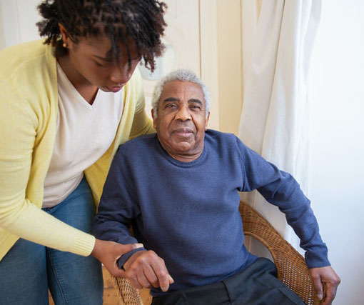 An elderly man sitting on a chair being assisted by a young lady.