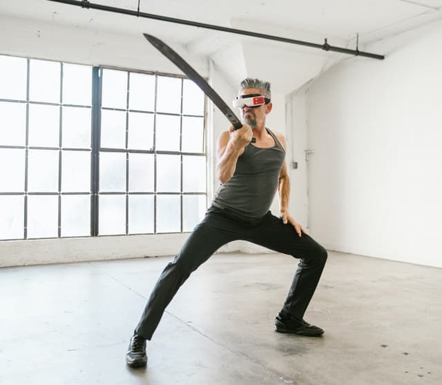 An elderly man wielding a sword and wearing a VR headset strikes a fighting pose.