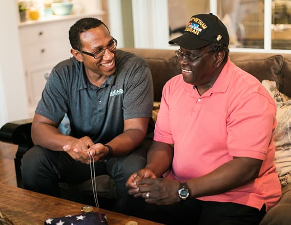 An older man and a man wearing an Amada shirt are conversing while laughing and seated next to each other on a couch.